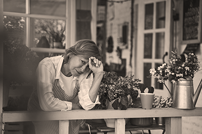 IMAGE: woman leaning on porch railing of rural house, hand to forehead appearing frustrated.