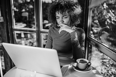 IMAGE: woman sitting at a table drinking coffee and working at a computer appears pensive.