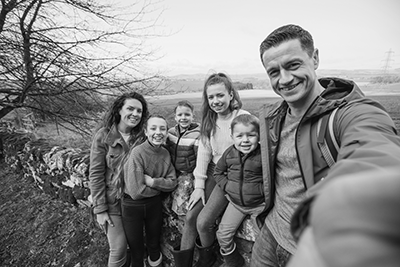 IMAGE: young family in a rural setting; woman, two girls, two boys, man all smiling in front of a stone wall.